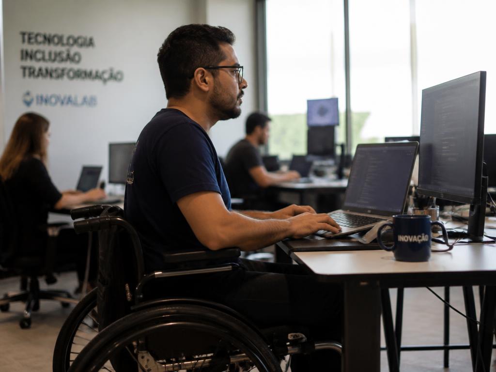 Profissional com deficiência usando cadeira de rodas trabalhando em estação de tecnologia em laboratório de inovação.